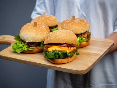 Lezzetli ev yapımı hamburger seti. Günlük beyaz tişört giyen bir kadın elinde dört lezzetli ev yapımı hamburger, tahta tepside ya da arka planda tahta kesme tahtası tutuyor..