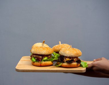 Lezzetli ev yapımı hamburger seti. El ele tutuşup, ahşap tepside ev yapımı dört hamburger ya da gri arka planda fotokopi alanı olan tahta kesme tahtası gösteriliyor..