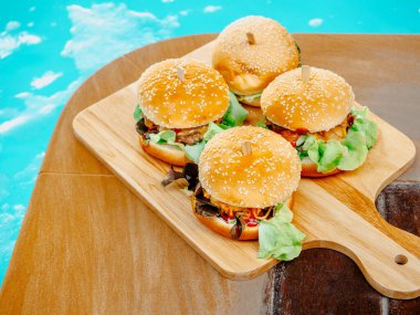 Delicious fresh homemade burger set. Four tasty homemade hamburgers on wooden tray or wood cutting board on dark background near the swimming pool.