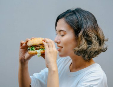 Delicious fresh homemade burger. Beautiful happy Asian woman short hair wearing casual white t-shirt holding and eating tasty homemade hamburger isolated on grey background.