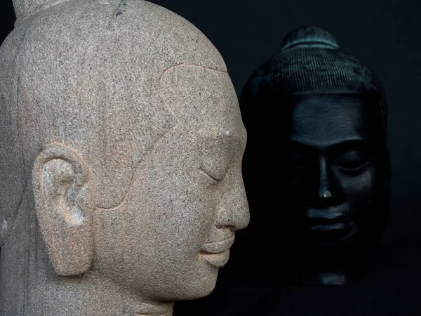 Two heads of Buddha carved from stone on dark background with copy space. The face of antique stone buddha, side view, and front view, close up.