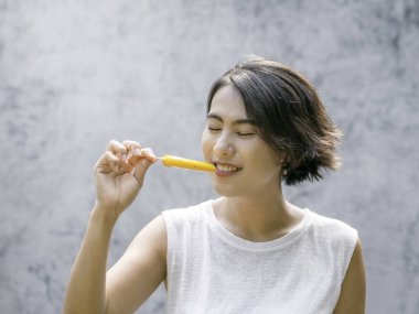 Dondurma yiyen bir kadın. Beyaz kolsuz tişört giyen mutlu, güzel Asyalı kadın açık havada, sarı buzlu şeker tutuyor. Gülümseyen kadın yazın buz lolipopunun tadını çıkarıyor..