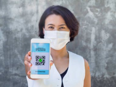 Uluslararası aşı sertifikası, akıllı dijital pasaport. Akıllı telefon ekranında QR kodu var. Omzunda yüz maskesi ve turuncu bandaj takan Asyalı bir kadın tarafından aşılanmış..