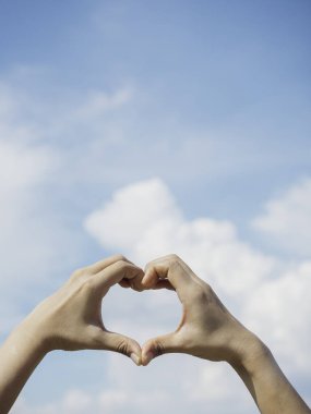 Mavi gökyüzünde kalp şeklinde eller ve tüylü bulut arkaplanı, aşk konsepti, ilişki ve romantik, dikey tarz. Kadın eli kopyalama alanı ile parmak sevgisi işareti yapıyor.