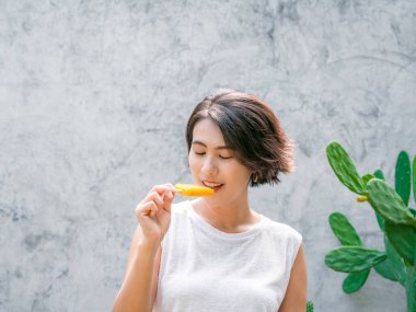 Dondurma yiyen bir kadın. Asyalı güzel, kısa saçlı, günlük beyaz kolsuz tişörtlü, açık havada sarı buzlu şeker tutan mutlu bir kadın. Gülümseyen kadın yazın buz lolipopunun tadını çıkarıyor..