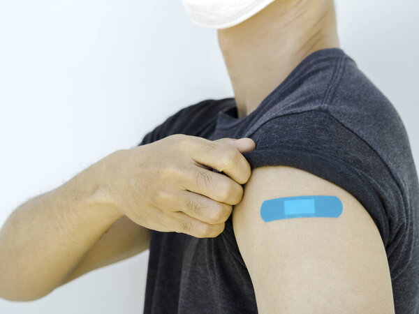 Vaccinations, bandage plaster on vaccinated people's arm concept. Blue color adhesive bandage on man's arm who wearing white face mask, showing own shoulder after vaccination treatment.