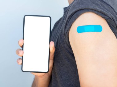 Sahte telefon, beyaz beyaz ekran akıllı telefondan tıbbi yüz maskesi takan ve omzunda mavi bandaj bandı olan beyaz fırında izole edilmiş bir adam tarafından gösteriliyor..