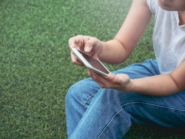 Cep telefonu, beyaz kolsuz gömlek ve mavi kot pantolon giyen, yeşil yapay çimlerin üzerinde oturan ve fotokopi alanı olan bir kadının elinde. Akıllı telefon tutan eller.