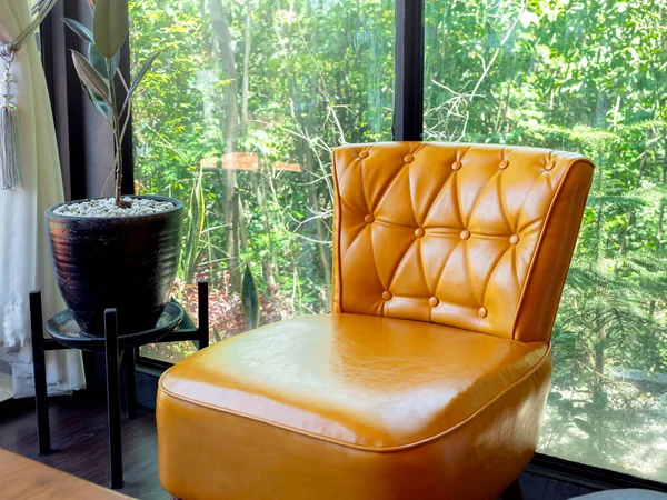 Yellow vintage leather sofa seat with pins and buttons decoration near plant pot and glass window with green nature view outside.