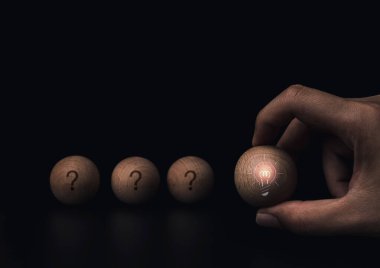 Fikir üretmek ve yaratıcı düşünceler üretmek. Parlak ışık fikri olan ahşap topu tutan el. Ampul ikonu. Koyu arkaplanda başka bir topun üzerinde soru işareti olan fotokopi alanı..