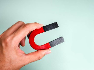 El ele tutuşan kırmızı ve siyah at nalı mıknatısı pastel yeşil arka planda izole edilmiş, cazibeyi, müşteri bağlılığını, kişisel markalaşmayı ve iş ve pazarlama kavramlarındaki manyetizmayı sembolize ediyor..