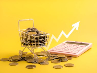 Hesap makineli bozuk paralarla dolu mini alışveriş arabası ve parlak sarı arka planda yukarı doğru ok. Yaşam, harcama kontrolü, enflasyon, bütçe planlaması ve mali yönetim kavramının artan maliyeti.