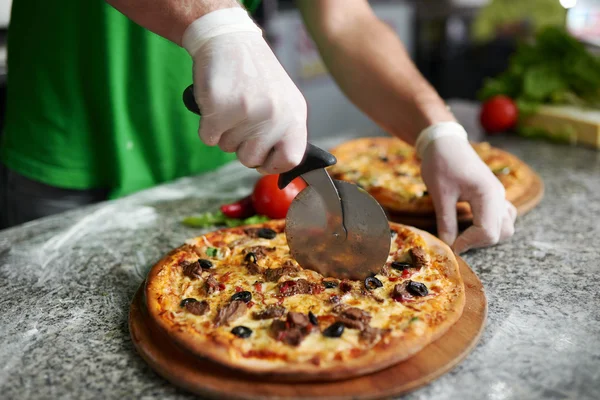 Chef cuts freshly prepared pizza on a wooden substrate.