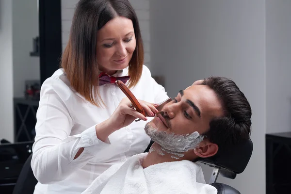 The barber woman shaves beard with razor and shaving foam