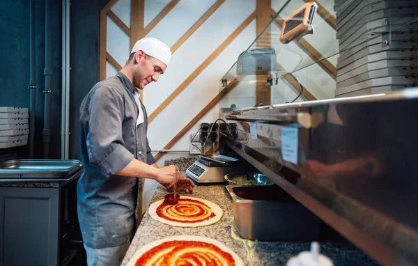 Een blij chef-kok strooit de saus op het pizzadeeg. Keukenwerk voor catering. — Stockfoto