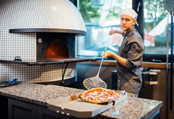 Chef doet vers gebakken hete pizza uit een spatel in een doos. Keukenwerk voor catering. — Stockfoto