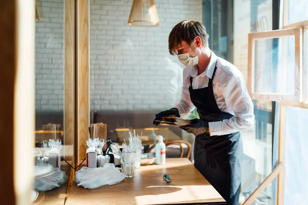 Un camarero alegre en una máscara está cerca de la mesa con productos de higiene personal y utensilios desechables. Precauciones durante la cuarentena en un café. Imagen de stock