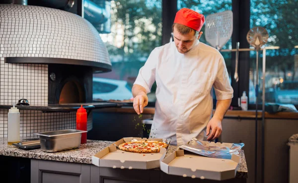 Szef kuchni posypuje pizzę w pudełku z przyprawami. Kuchnia cateringowa. — Zdjęcie stockowe