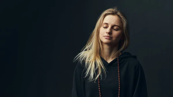 Mujer rubia con rosario en el cuello meditar sobre fondo negro. — Foto de Stock