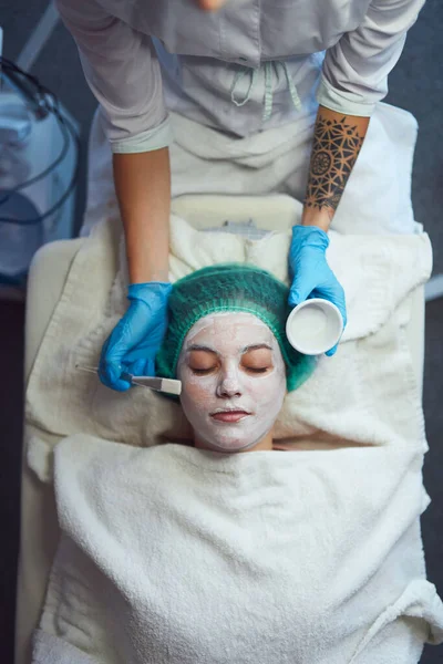 Косметолог наносит женщине терапевтическую маску на лицо. Женщина в спа-салоне по косметическим процедурам для ухода за лицом. — стоковое фото
