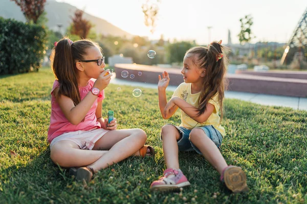 Two girls are sitting on the grass and blowing bubbles — Stock Photo, Image