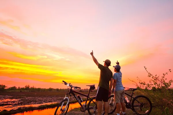 Bakifrån av en ung man som står med en cykel vid solnedgången. läka — Stockfoto