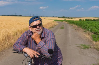 Sakallı, tombul, yaşlı bir adamın açık hava portresi. Kırsal bir yolda yaz tatilinde kısa dinlenirken bisikletinin barını tutmak için dirseklerini eğiyor.