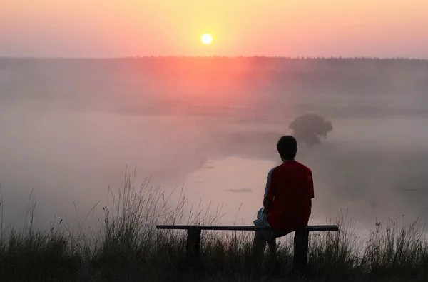 Hombre sentado en el banco y viendo el amanecer brumoso . — Foto de Stock