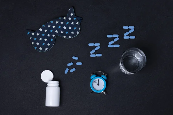 Beyaz şişe ve haplar, bir bardak su, mavi alarm saati ve siyah arka planda uyku maskesi. Uyku arkaplanı. Melatonin, uykusuzluk ve iyi geceler.