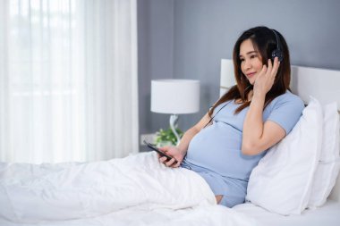 Hamile bir kadın yatakta kulaklıkla müzik dinlemek için akıllı telefon kullanıyor.