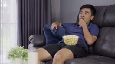 Oturma odasındaki kanepede otururken uzaktan kumandayı elinde tutan ve televizyon izleyen neşeli genç adam. 