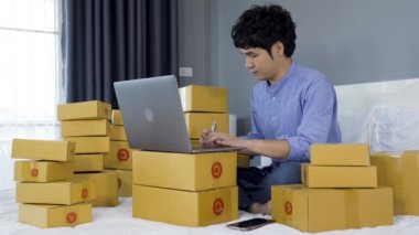 Genç bir adam evdeki bir yatakta online ürün satmak için dizüstü bilgisayar kullanıyor ve çalışıyor.