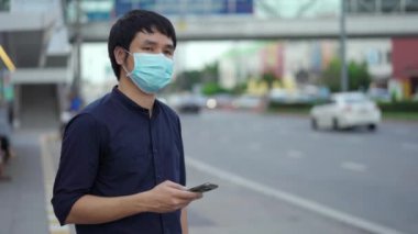 Asyalı genç adam akıllı telefon tutuyor ve otobüs durağında otobüs bekliyor ve Coronavirus (covid-19) salgınının yayılması için maske takıyor.