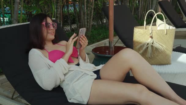 ビキニ姿の若い女性がデッキチェアに寝そべってプールの近くのスマートフォンを使って — ストック動画