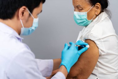 Doktor elinde şırınga tutarken ve pamuk kullanırken kıdemli bir hastaya tıbbi maske takıyor. Covid-19 veya koronavirüs aşısı