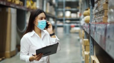Tıbbi maskeli kadın çalışan cep telefonuyla konuşuyor ve Coronavirus (covid-19) salgını sırasında depodaki stokları kontrol etmek için pano tutuyor..