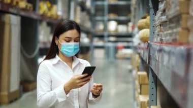 Coronavirus (covid-19) salgını sırasında depodaki envanteri kontrol etmek için akıllı telefon kullanan tıbbi maskeli kadın çalışan..