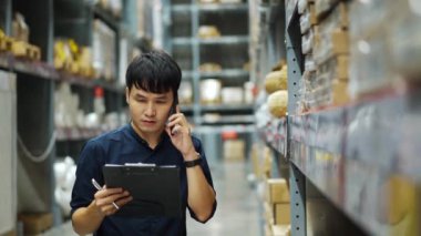 Depodaki envanterleri kontrol etmek için cep telefonuyla konuşan ve elinde bir pano tutan genç adam.