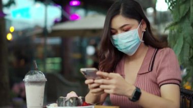 Kafedeki Coronavirus (covid-19) salgınından korunmak için akıllı telefon kullanan ve tıbbi maske takan kadın