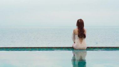 Yüzme havuzunun kenarında oturan ve denize bakan genç bir kadın.