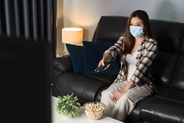 Coronavirus 'u korumak için kanepede televizyon izleyen ve tıbbi maske takan genç bir kadın (Covid-19)