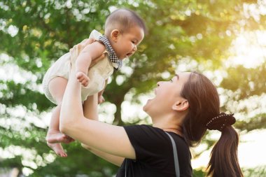 Anne kucağında yeni doğmuş bir bebeği kollarıyla kaldırıyor. Yeşil bir arka planda, güneş ışığıyla birlikte yükseliyor.