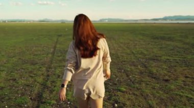 Neşeli kadın yeşil arazide gün batımıyla yürüyor.