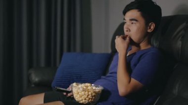 Genç adam kanepede oturup gece televizyon izlerken patlamış mısır yiyor.
