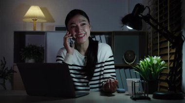 Bir gece dizüstü bilgisayarla çalışırken akıllı telefonla konuşan mutlu bir kadın.