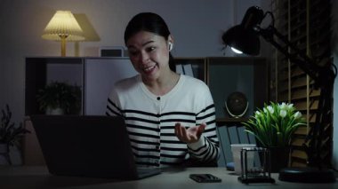 Diz üstü bilgisayar ve kablosuz kulaklık kullanan mutlu bir kadın bir gecede video görüşmesi yapıyor.