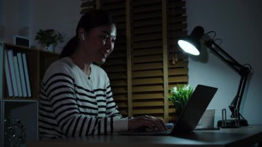 Diz üstü bilgisayar ve kablosuz kulaklık kullanan mutlu bir kadın bir gecede video görüşmesi yapıyor.