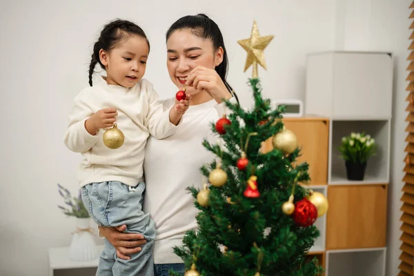 Mutlu anne ve küçük kız Noel ağacını evde oyuncak topla süslüyorlar.