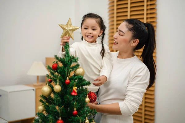 Mutlu anne ve küçük kız evdeki Noel ağacında süsleme yapıyor.