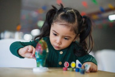 Çocuk kız masaya alçı heykel boyuyor.